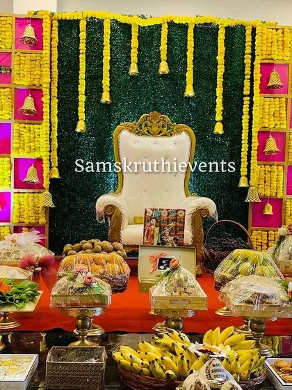 A full view of a festive Seemantha setup, highlighting the wrapped offering plates and fruit baskets. The bright yellow and pink backdrop with hanging bells adds to the celebratory atmosphere.