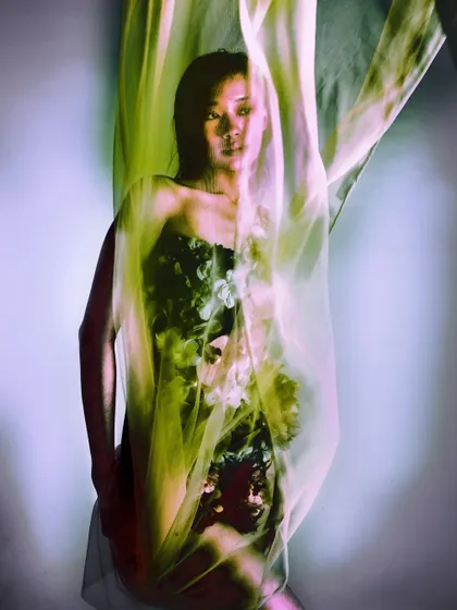 The model is partially obscured by a sheer, flowing veil of fabric, creating a sense of mystery and distance. The soft, greenish light adds to the underwater, dreamlike atmosphere.