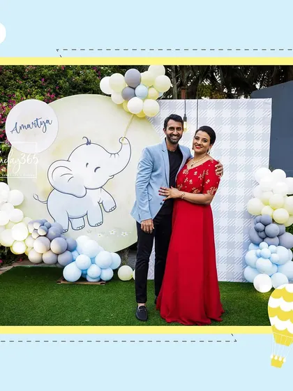 A lovely photo of the happy parents at their son's baby elephant-themed celebration. They are posing in front of a custom backdrop featuring a cute elephant illustration and soft balloon clouds.