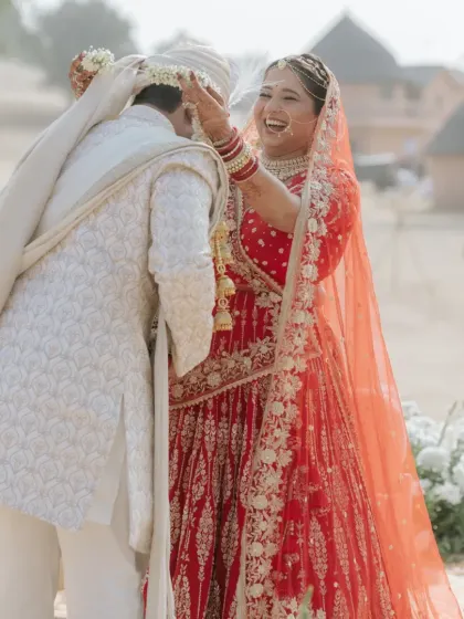 The groom playfully helps the bride with her turban during a lighthearted moment in their wedding ceremony.