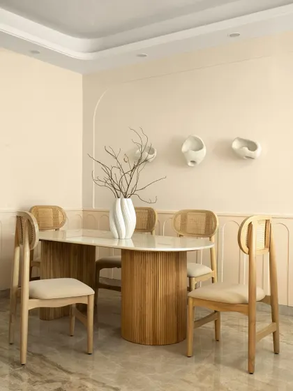 The dining area of Casa Levante embraces a Wabi-Sabi elegance. A fluted wooden table base is topped with marble, surrounded by cane-back chairs. The wall is adorned with minimalist ceramic art.