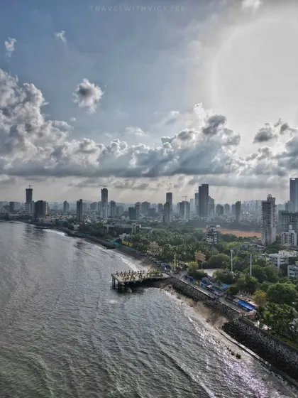 A wide drone shot of the Dadar coastline, capturing the viewing deck, the sea, and the expansive Mumbai skyline under a dramatic, sunlit sky.