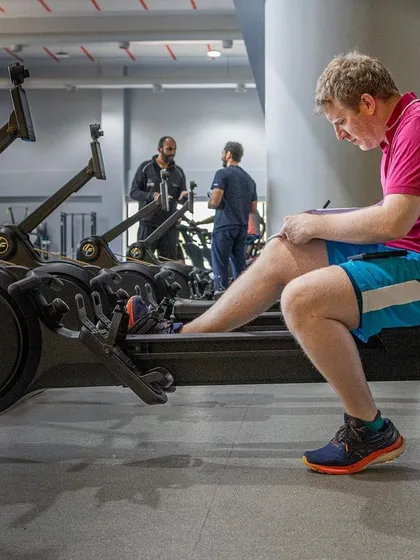 A UKSCA trainer takes notes while observing a participant on a rowing machine, assessing their conditioning and technique.