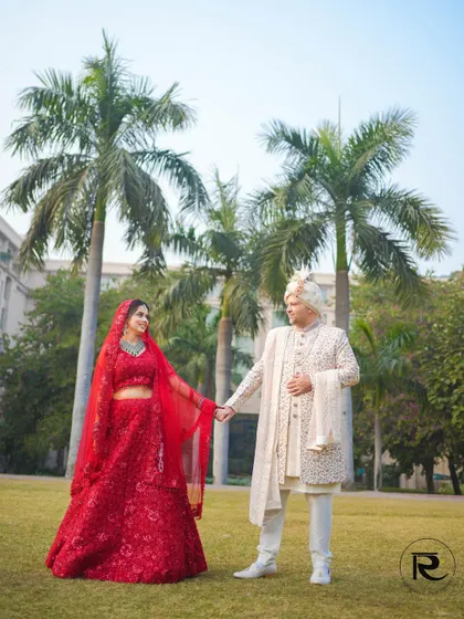 A classic wedding day portrait. The couple holds hands in a beautiful garden, the bride's red lehenga vibrant against the green backdrop.