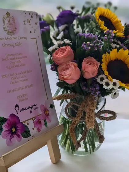 The custom menu for the Varnya salon launch, surrounded by a beautiful bouquet of sunflowers and roses. The menu details the gourmet tacos, sliders, and charcuterie board we prepared.
