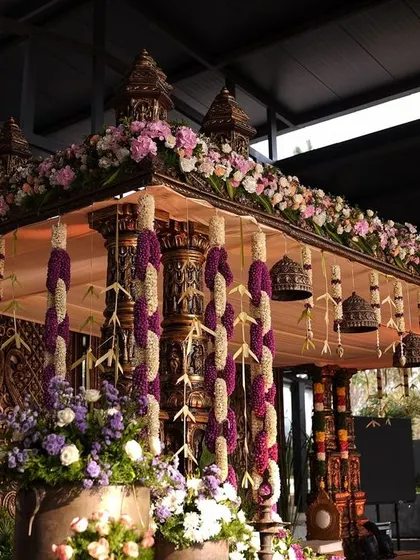 A grand South Indian mandap featuring intricately carved pillars adorned with purple and white floral garlands and traditional temple bells.