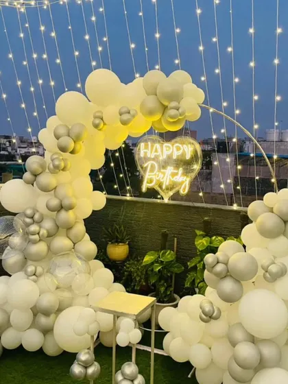 A chic rooftop birthday party with a white and silver theme. This setup features a balloon half-arch under a canopy of fairy lights and a heart-shaped neon sign.