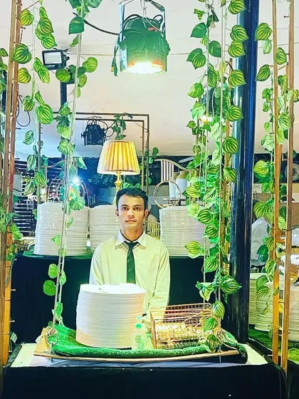 A member of our team ready to serve at our green-themed buffet station. The setup is designed to be both beautiful and functional, with stacked plates and elegant lamps.