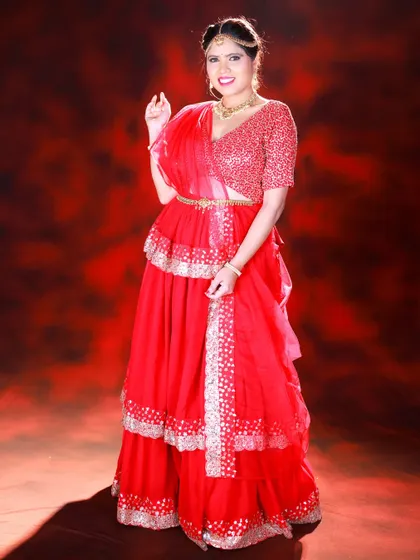 A full-length studio portrait against a dramatic red background. The lighting highlights the sequin work on the blouse and the tiered layers of the skirt.