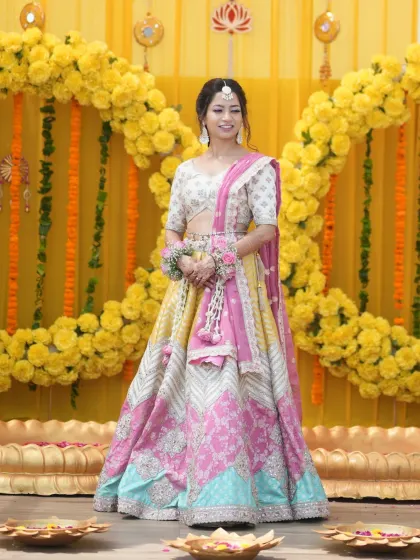 A full-length shot of the bride against a marigold backdrop. Her makeup is soft and elegant, complementing her beautiful multi-colored lehenga.