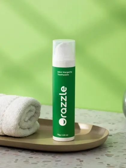 A simple lifestyle scene for Orazzle toothpaste, set in a bathroom environment. The terrazzo tile, rolled towel, and small plant create a realistic and aesthetically pleasing context for the product.