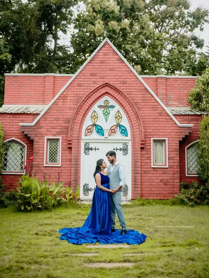 A wide shot of a couple standing before a picturesque red chapel, with the mother-to-be's long blue gown spread out on the grass, creating a dramatic and romantic portrait.