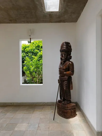 A window in a simple white corridor frames the lush green foliage outside, turning the view into a living piece of art. A traditional wooden sculpture stands guard, connecting the space to its cultural roots.
