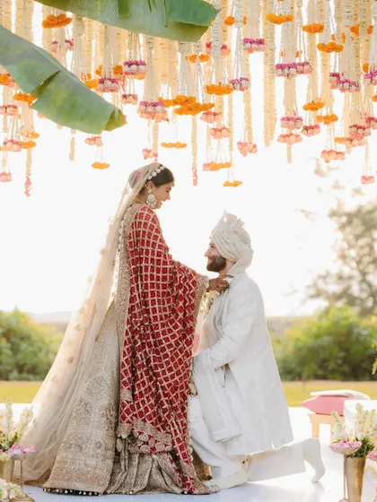 A tender moment during the varmala ceremony where the groom is on one knee before his bride under a canopy of flowers.