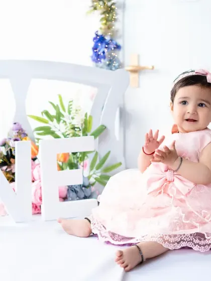 Clapping with joy, this little girl celebrates turning one. The "ONE" sign is a classic prop for first birthday photoshoots, marking this important milestone.