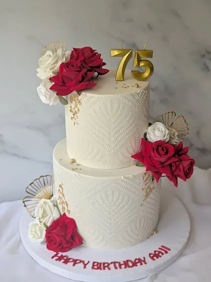 Celebrating 75 years of Aaji's grace. This two-tier cake features a delicate white-on-white stencil pattern, beautifully contrasted with deep red and white roses and elegant gold fans.
