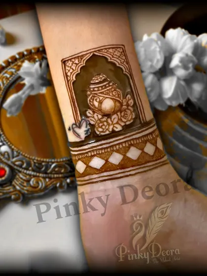 A simple and auspicious design for an engagement ceremony. This wrist mehndi features a traditional kalash (pot) in a jharokha (window), symbolizing prosperity and new beginnings.