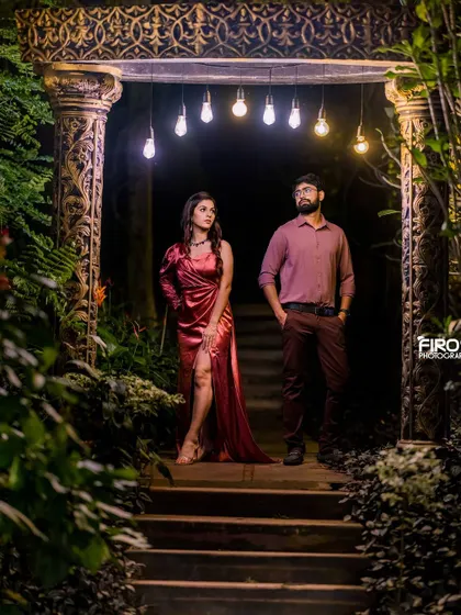 A dramatic and moody shot of a couple on a staircase, framed by lush greenery and lit by hanging bulbs.