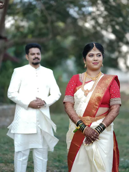 A formal portrait of a bride and groom in an outdoor setting. The bride stands in the foreground in her elegant saree, while the groom stands behind, creating a classic and composed wedding photograph.