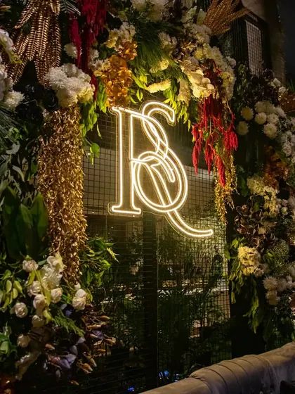 A custom neon monogram serves as a modern and personalized photo backdrop. It is framed by an extravagant arch of flowers and foliage, blending contemporary style with natural beauty.