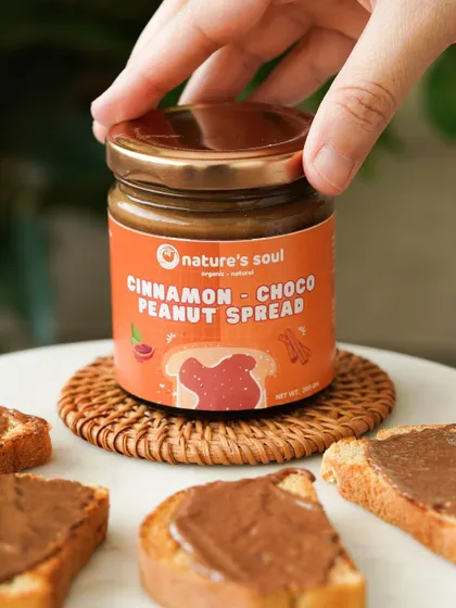 A hand opening a jar of cinnamon choco peanut spread, surrounded by toast points, creating a sense of anticipation.