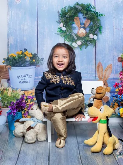 A happy little boy in a traditional outfit, sitting in a spring-themed set filled with bunnies, flowers, and a stuffed Pluto. His joyful shout is a perfect candid moment.