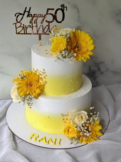 A sunny and cheerful two-tier cake for a 50th birthday party. The yellow ombre design is decorated with beautiful fresh yellow gerberas and baby's breath.