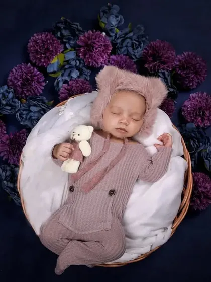 A baby in a cute bear-ear romper holds a tiny teddy bear, surrounded by a wreath of purple flowers against a deep blue background.