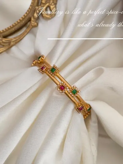 A close-up of a gemstone bangle from Manjali Jewellers, draped in soft white cloth. This styling focuses on the texture of the fabric and the sparkle of the stones, creating a feeling of pure luxury.