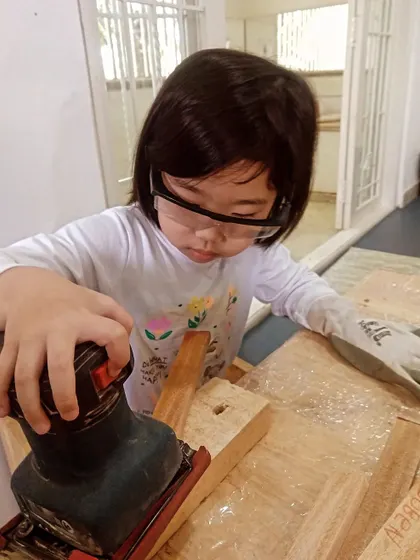 A young innovator learns the importance of preparation, using a power sander to smooth the edges of her wooden project. It's the first step to a professional finish.