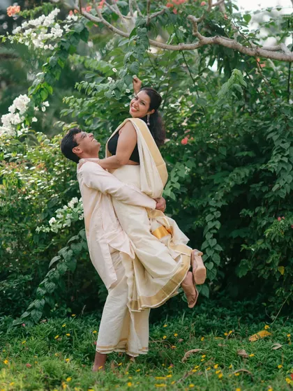 A playful and romantic moment as the groom lifts the bride, both dressed in elegant South Indian attire amidst a lush garden.