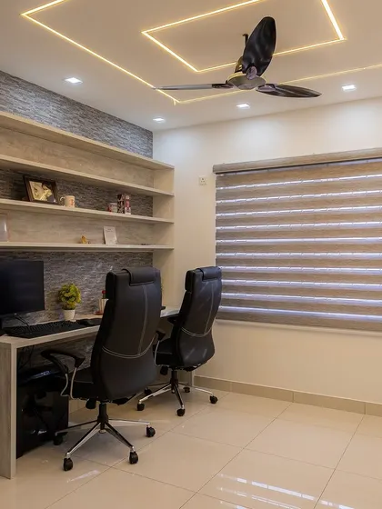 This home office is designed for two, with a long desk and ergonomic chairs. The textured wall and floating shelves add style, while the false ceiling with profile lighting provides excellent illumination.