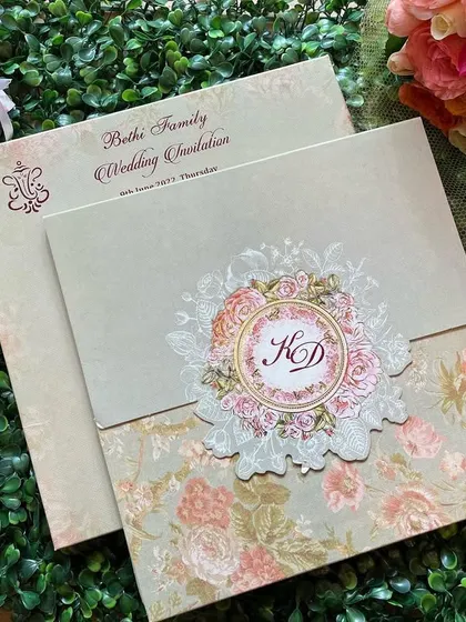 A sophisticated invitation with a vellum overlay featuring a floral wreath monogram. The pastel green and pink color scheme is soft and romantic.