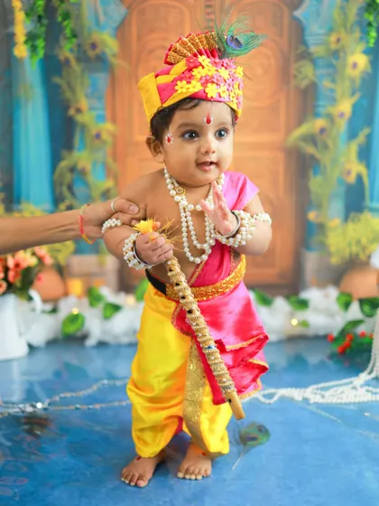 A little Kanha standing with his flute, looking like a true divine prince. The hand of a parent offering support just out of frame is a reminder of the love surrounding him.