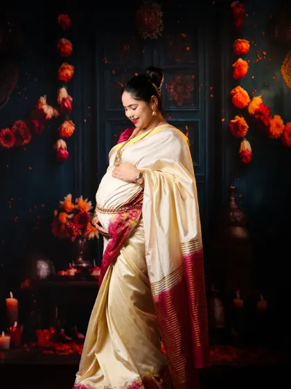 A graceful solo portrait celebrating tradition. The mom-to-be looks radiant in a classic cream and red saree, standing against a festive backdrop of flowers and candles.