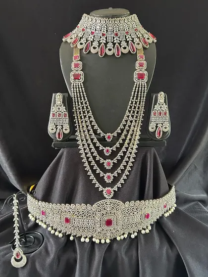 For a touch of colour, this silver-toned set with ruby-red stones is a perfect choice. The full set includes a choker, a layered necklace, and a matching vaddanam for a complete party look.