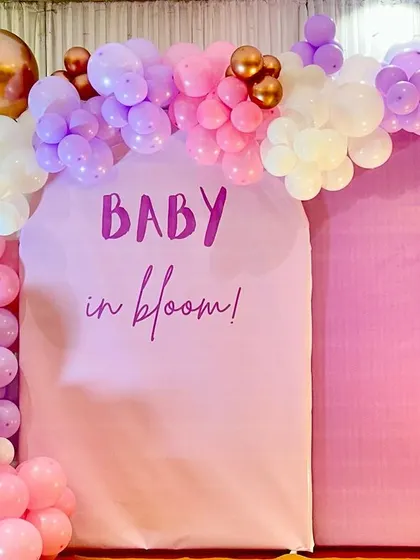 A "Baby in Bloom" themed baby shower backdrop. The pink arches are decorated with a beautiful balloon garland in pastel pink, purple, and gold.