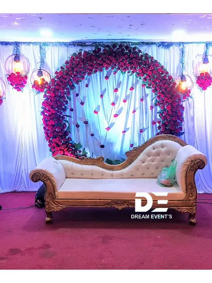 This romantic ring ceremony stage features a large circular arch of red roses against a soft white curtain. Hanging glass lanterns with flowers inside add a touch of whimsy and magical lighting to the decor.