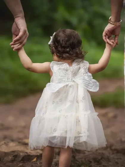 A toddler in a delicate white dress with butterfly wings holds her parents' hands as they walk through a natural path. This shot, focusing on their linked hands, symbolizes guidance, love, and the journey of family.