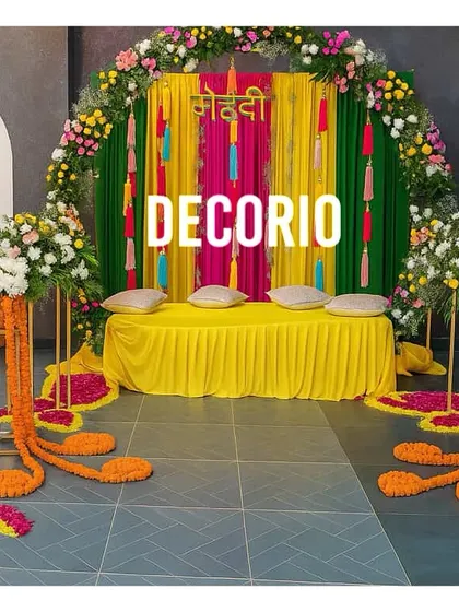 A colorful Mehndi setup featuring a backdrop of rich green and pink drapes. The comfortable floor seating with bright yellow fabric and cushions provides a perfect spot for the bride during the ceremony.