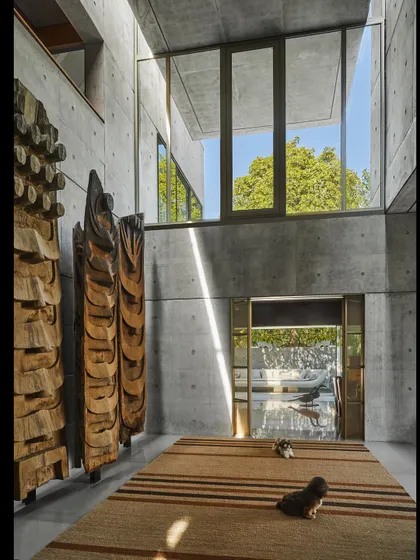 The entrance vestibule of a residence in Delhi, featuring fair-faced concrete walls and a double-height window. The space is animated by the play of sunlight and the inclusion of large-scale wooden sculptures.