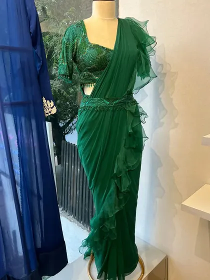 A studio view of our emerald green pre-draped saree. Crafted from pure georgette with hand embroidery and ruffles, it comes with a matching blouse and waist belt for a complete cocktail look.