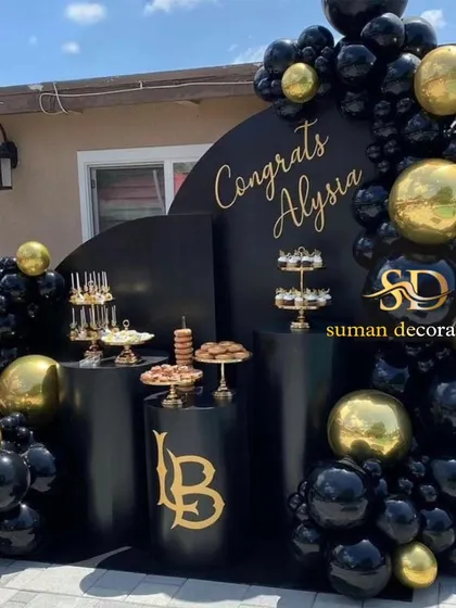 A sophisticated black and gold setup for a graduation party. This includes a custom arched backdrop, black plinths with custom decals, and a balloon garland.