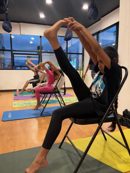 A young student demonstrates incredible focus and flexibility, using a chair to support a standing leg-to-head pose.