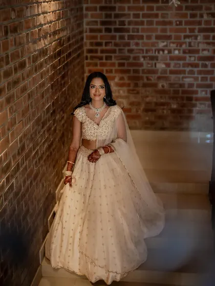 A full-length shot of the bride on a staircase. The soft, elegant makeup balances the grandeur of her white lehenga, creating a look of timeless grace.