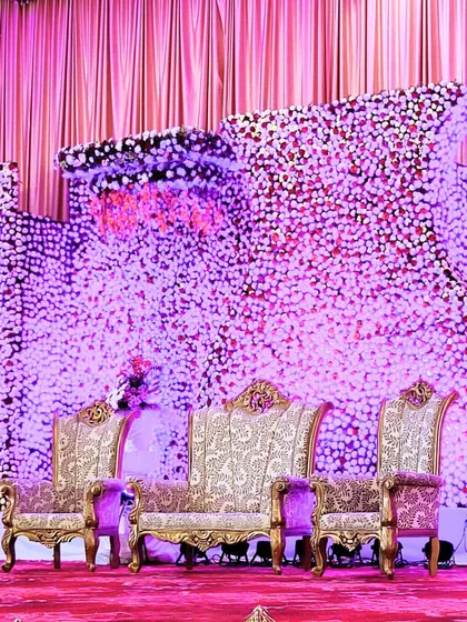 A regal reception stage with seating upholstered in rich brocade fabric. The backdrop is a dense wall of purple and white flowers, creating a luxurious and opulent setting.