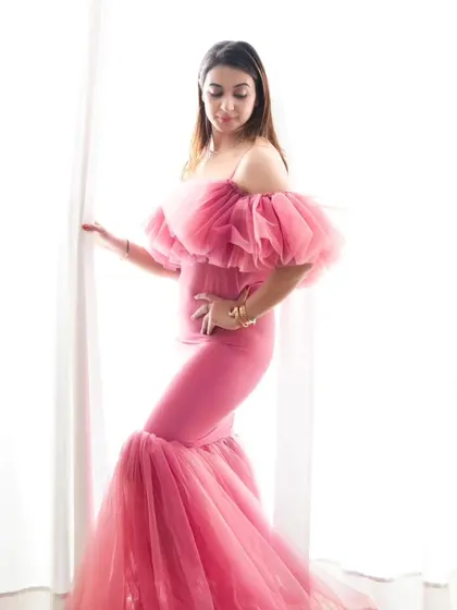 A high-key portrait that feels ethereal and light. The mother-to-be, dressed in a dramatic pink tulle gown, is silhouetted against a bright window, creating a soft, dreamy effect.