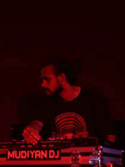 A moody, red-lit shot of me at the DJ booth, with my Theyyam-inspired logo visible on my equipment.
