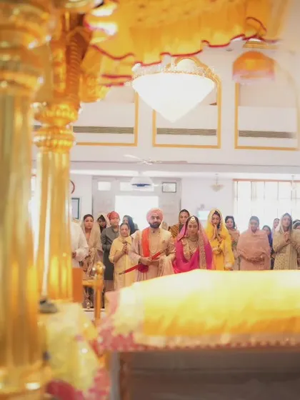 The couple stands before the Guru Granth Sahib during their Sikh wedding. This wide shot captures the entire scene, with family looking on in support.
