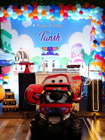 Another view of the car theme decoration for Tansh's birthday. This setup features a toy monster truck, character cutouts, and a vibrant balloon garland, making it a dream party for any little car enthusiast.